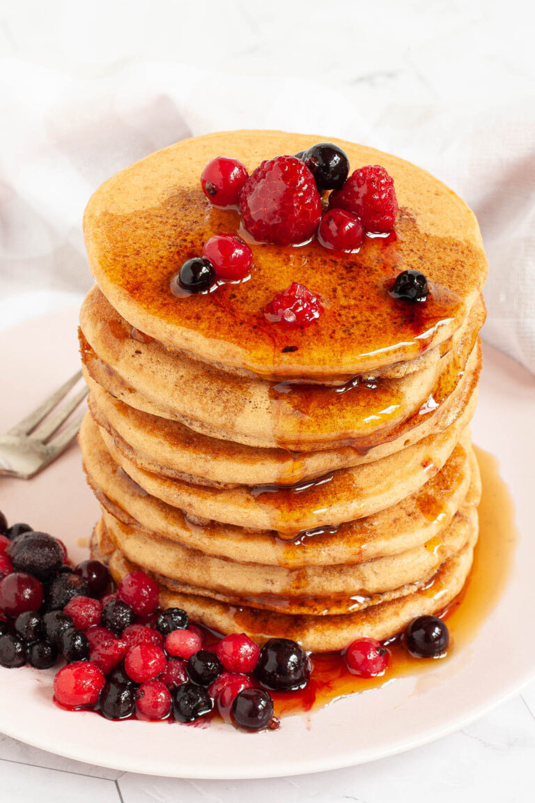 Pink plate with a stack of pancakes topped with black and red berries and maple syrup