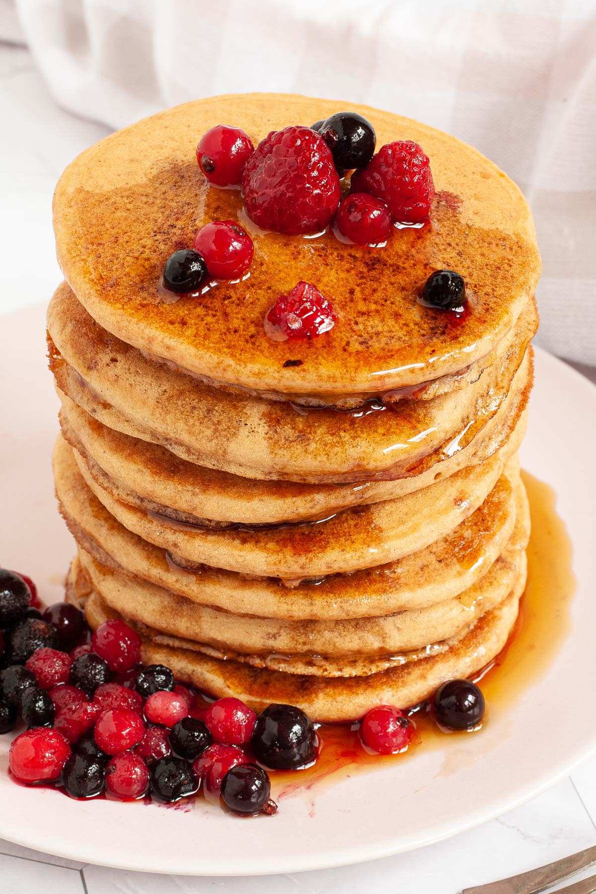 Pink plate with a stack of pancakes topped with black and red berries and maple syrup