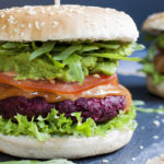 Close-up of a beet burger with layers like burger bun, lettuce, purple patty, tomato slice, green puree, arugula and bun on the top