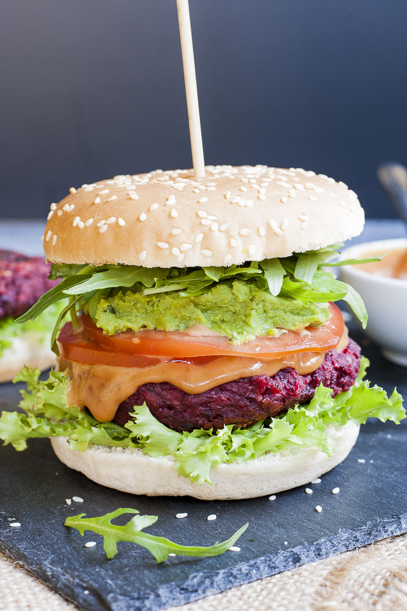 Close-up of a beet burger with layers like burger bun, lettuce, purple patty, tomato slice, green puree, arugula and bun on the top