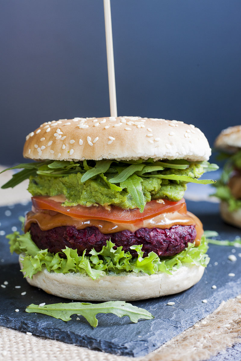 Close-up of a beet burger with layers like burger bun, lettuce, purple patty, tomato slice, green puree, arugula and bun on the top
