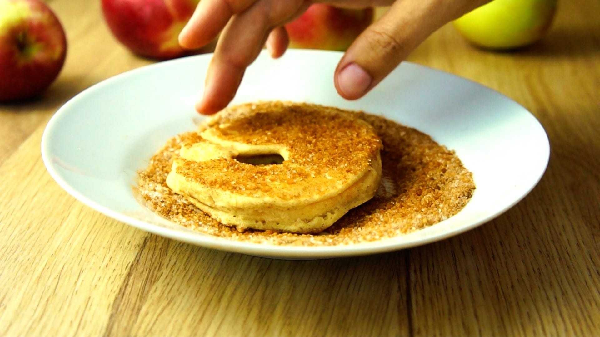 White plate sprinkled with cinnamon sugar and a fried apple ring is coated using a hand. 