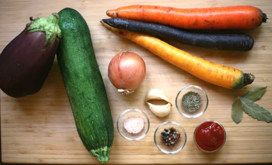 The ingredients are placed on a wooden board: carrots, zucchini, eggplant, onion, garlic cloves, small glass of salt, black pepper, tomato paste and thyme
