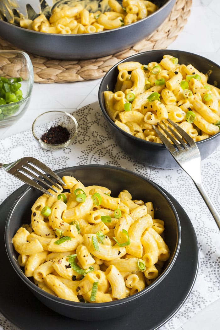 2 black bowls of pasta in creamy yellow sauce topped with chili flakes and chopped green onion. Remaining pasta is in a frying pan in the corner. 