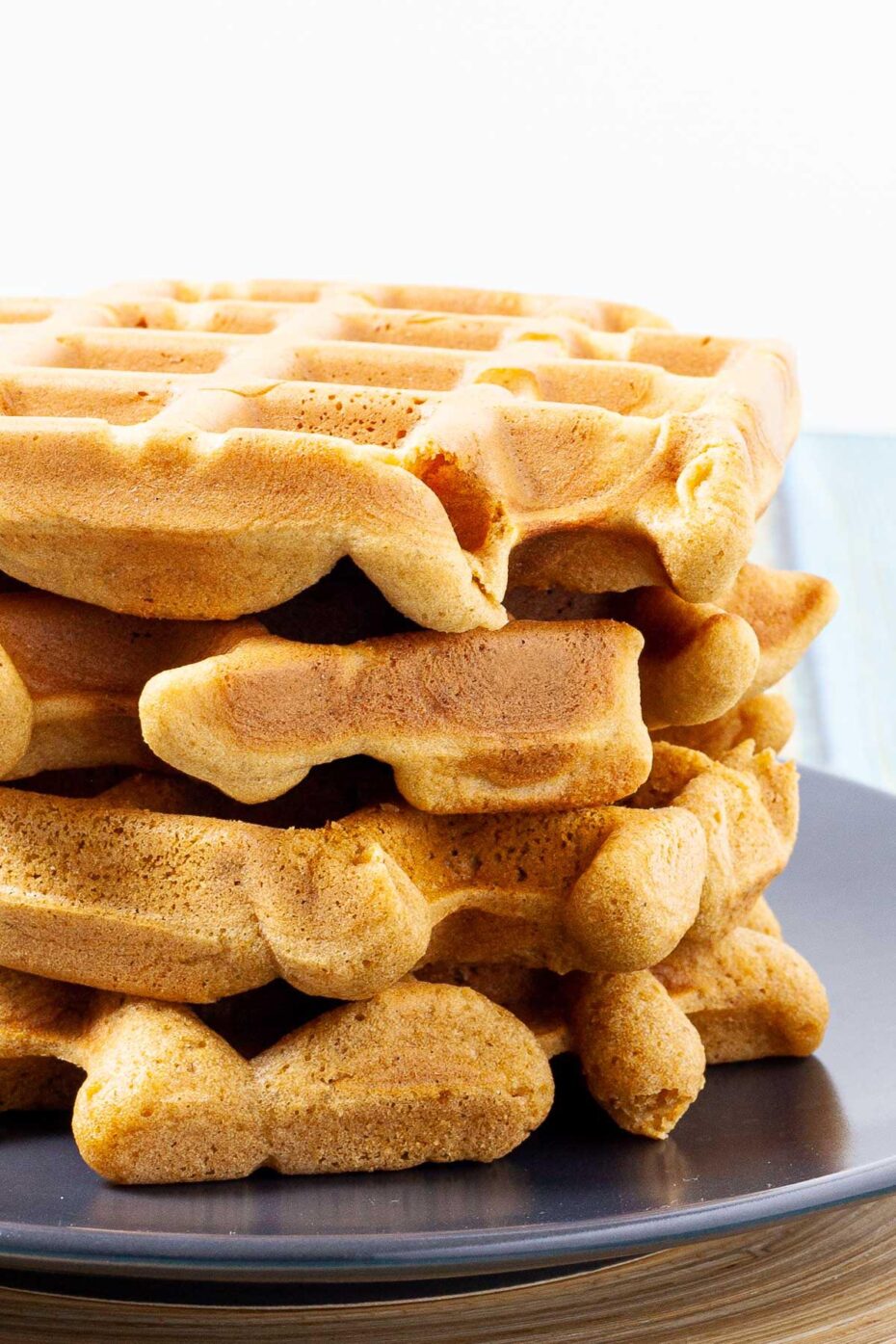 Black plate with a stack of waffles topped with sliced banana and maple syrup. There are 3 waffles on another black plate in the background.