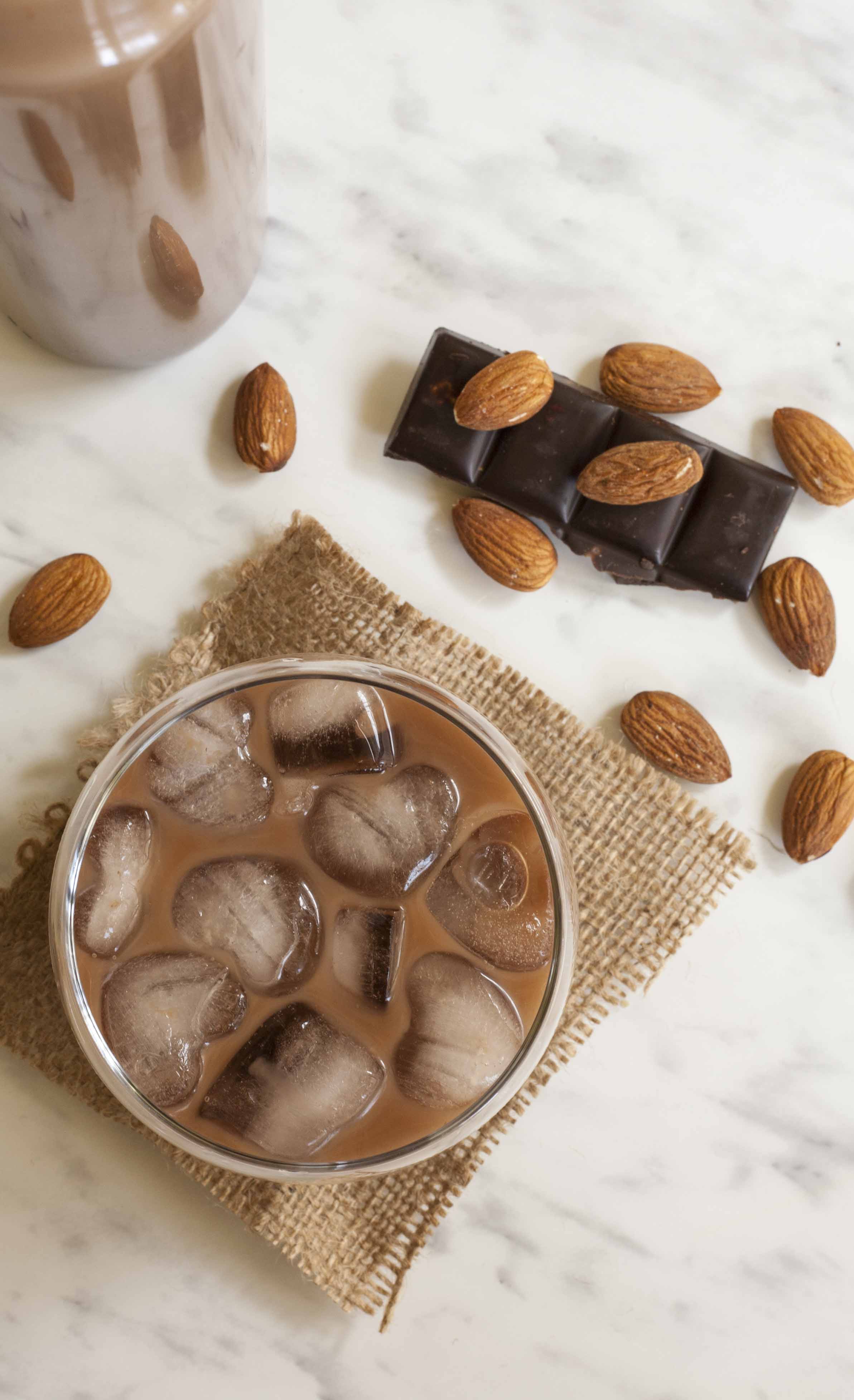 A whiskey glass with brown creamy drink and heart-shaped ice. Almonds, chocolate cubes are scattered around it. 