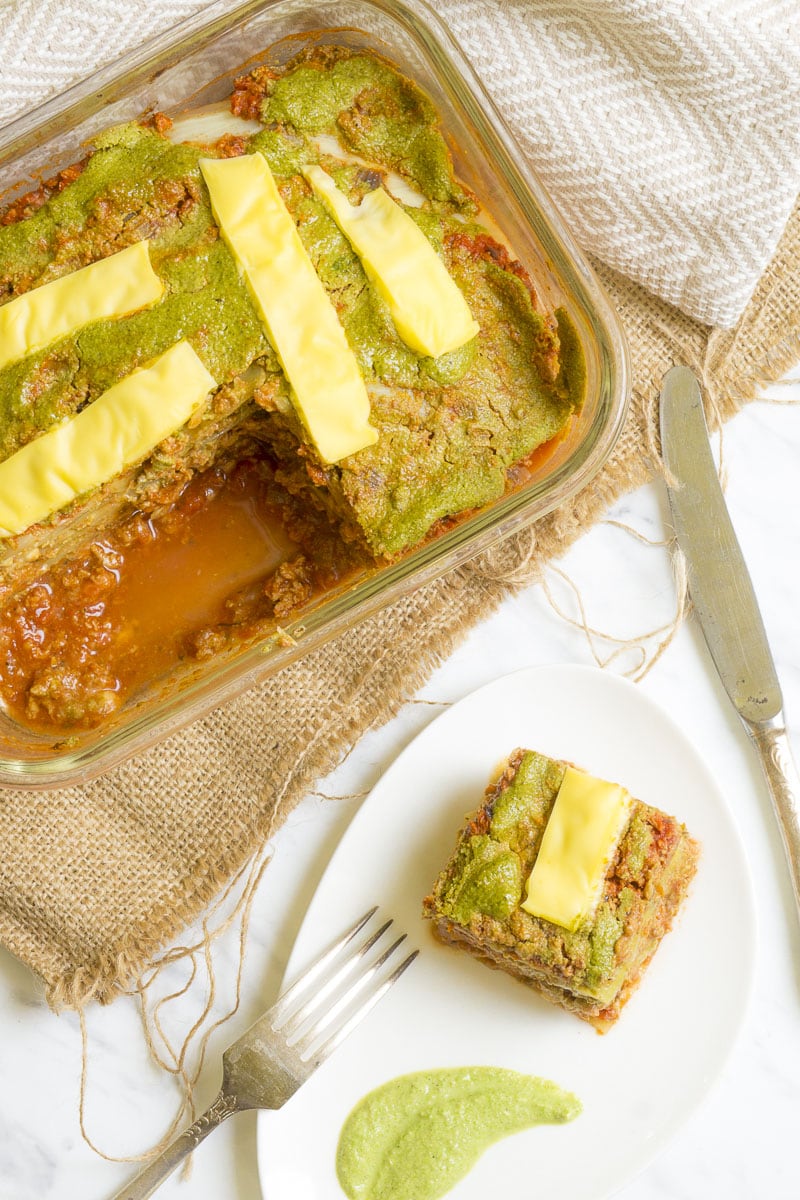 Glass pan full of vegan lasagna from above. Next to it a slice of lasagna on a white plate with a green spinach alfredo slushed in a shape of a leaf