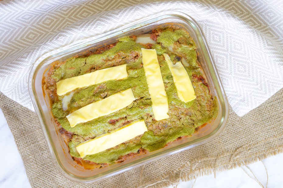 Glass pan full of vegan cabbage lasagna from above. Top layer of cheese slices and below green spinach alfredo is visible clearly