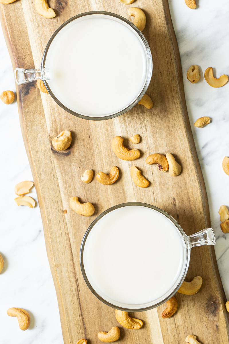 Two glass mugs on a wooden board shown from the top full of cashew milk. Cashew nuts sprinkled around.