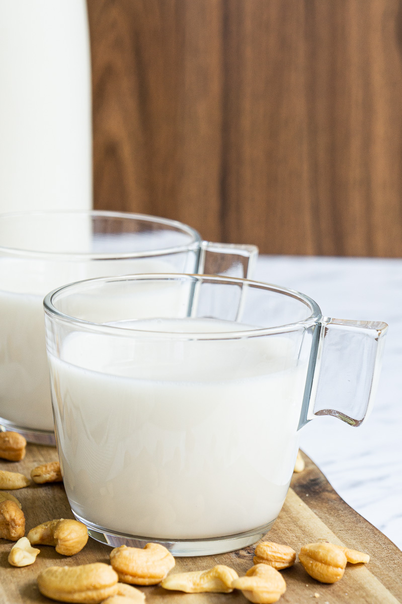 Glass mug on a wooden board full of cashew milk. Cashew nuts sprinkled around.