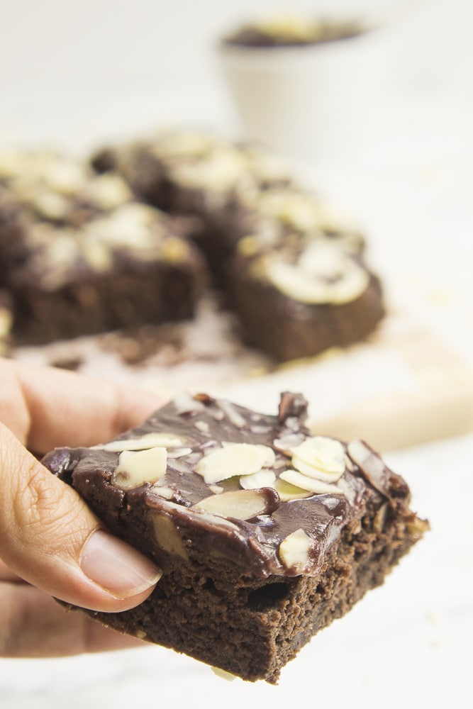 Hand holding one slice of vegan sweet potato brownies with chocolate frosting and sliced almonds. Other slices are in the background.