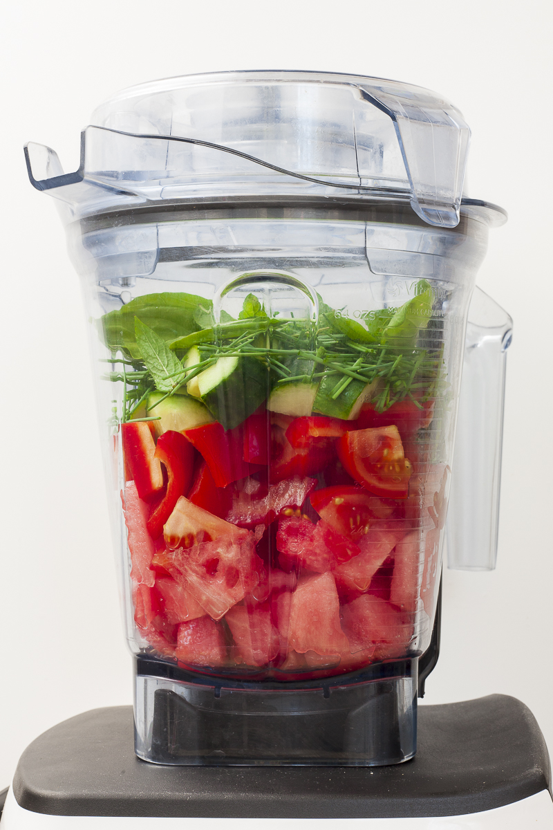 Blender from the side that contains raw red and green diced vegetable with herbs and green leaves on the top.
