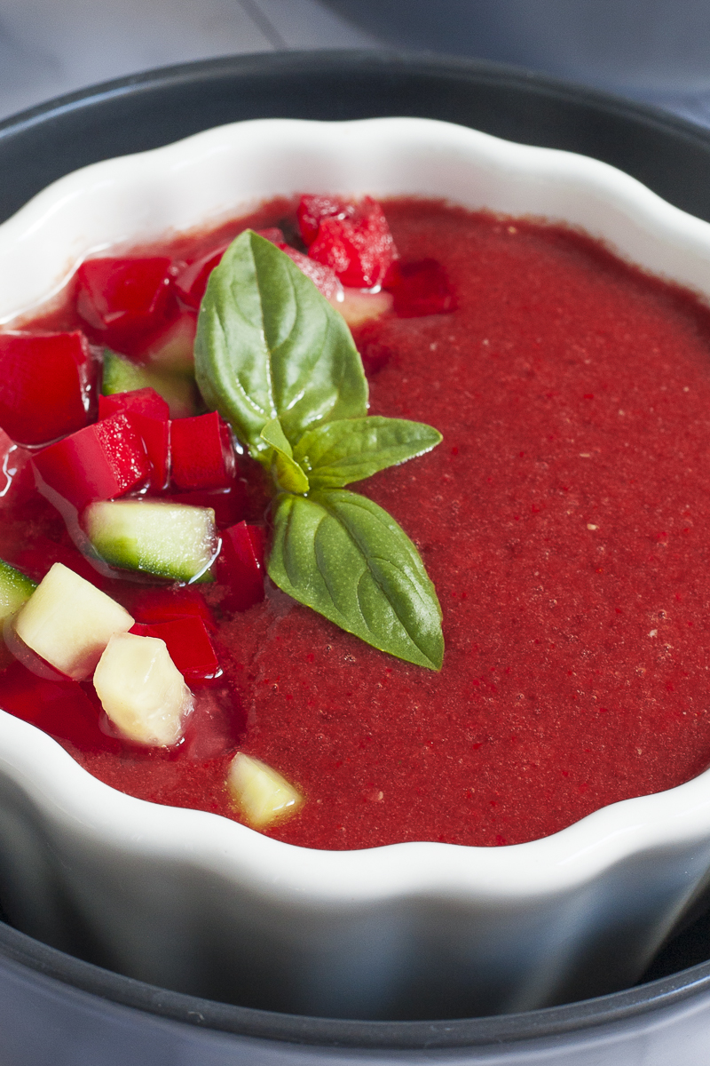 Red soup in a white and black bowl topped with basil leaves and diced red and green vegetables.