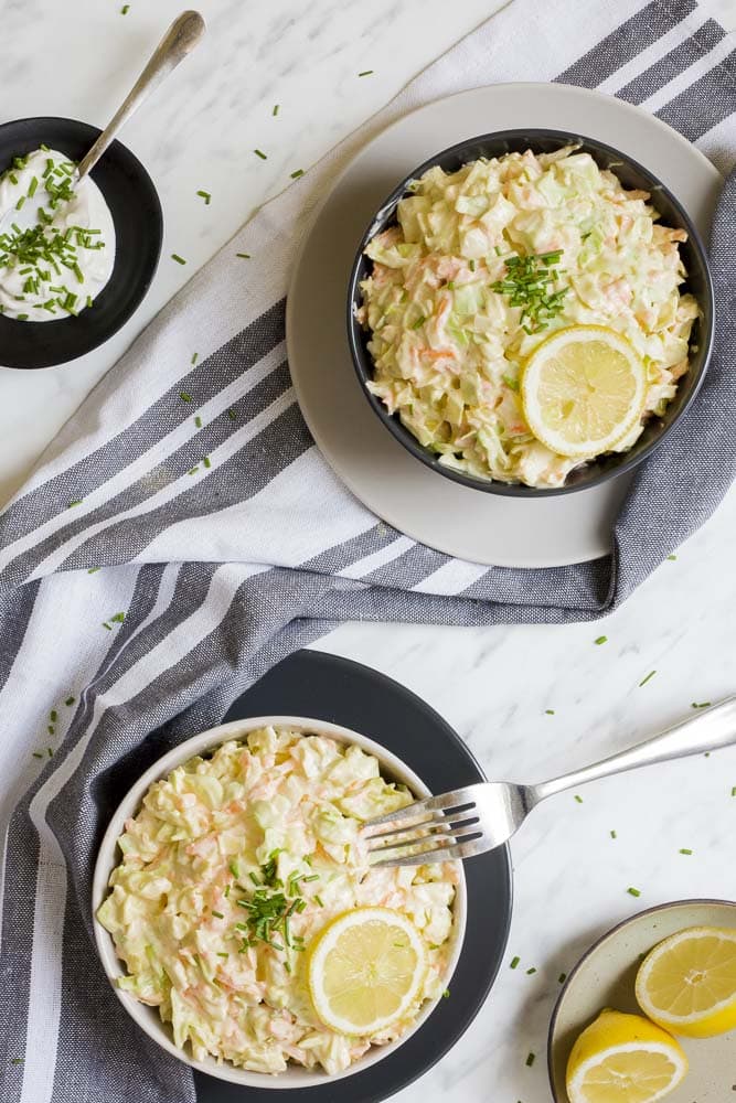 White bowl of shredded cabbage and carrots in a creamy sauce. A fork is taking a bite. The same in a black bowl is at the back. 