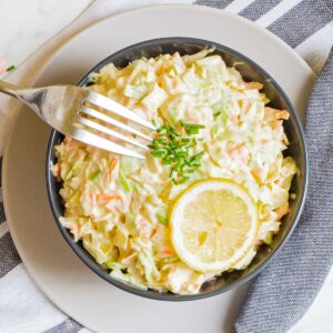 Black bowl with shredded cabbage and carrots in a creamy dressing topped with a lemon slice and some chopped chives.