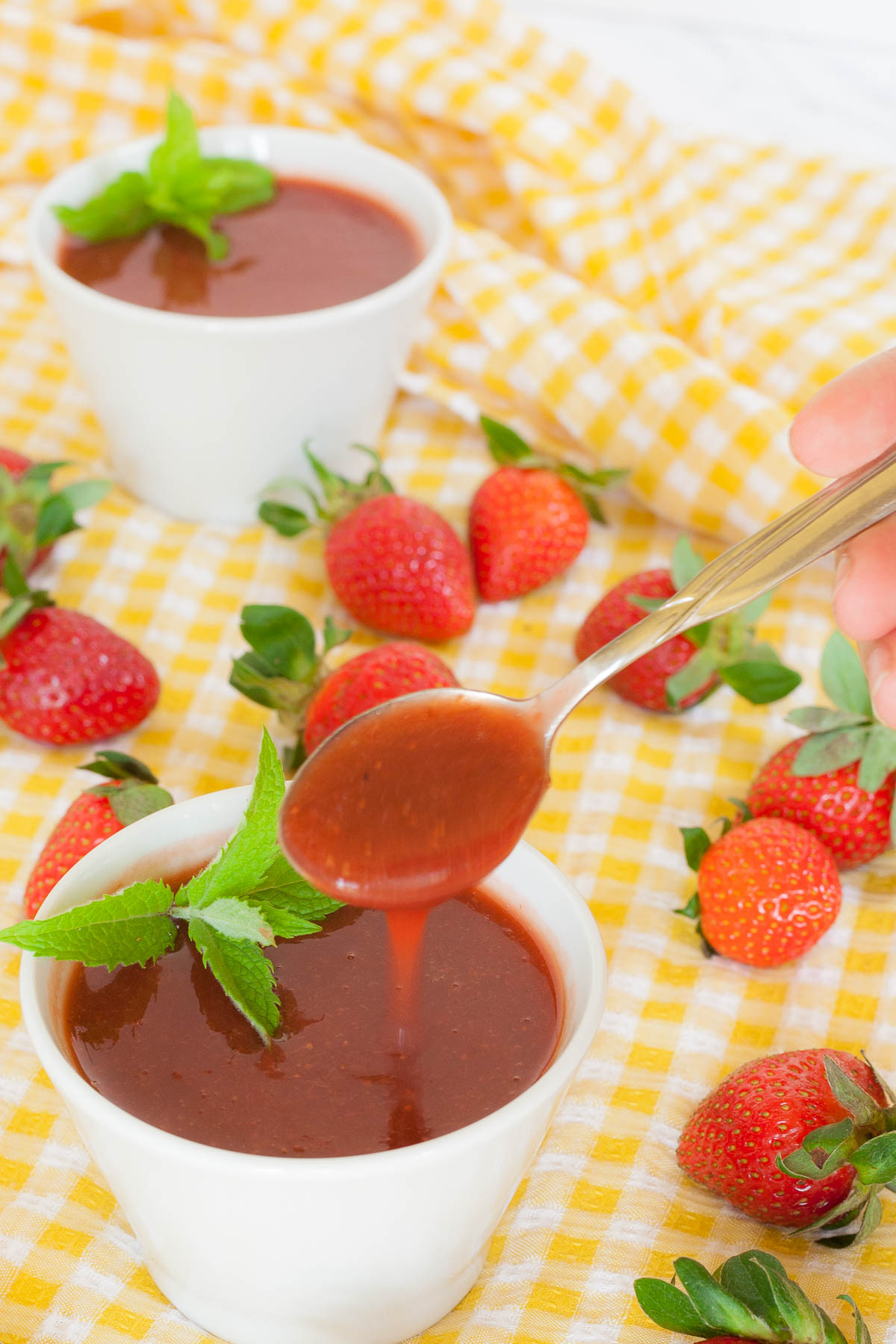 Small white bowl with strawberry puree and some mint leaves. More fresh strawberries are scattered around it on a yellow tablecloth. A hand is holding a spoon to take some.