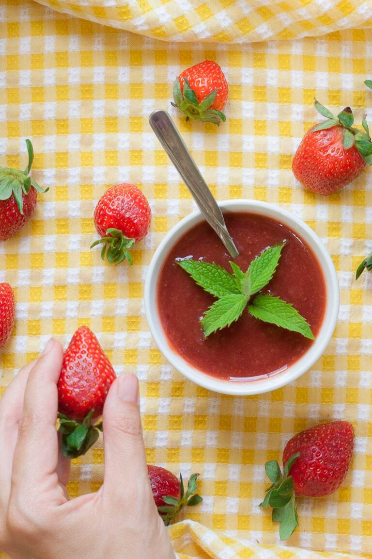 Two small white bowls with strawberry puree and some mint leaves. More fresh strawberries are scattered around it on a yellow tablecloth. A hand is holding one strawberry.