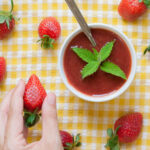 Two small white bowls with strawberry puree and some mint leaves. More fresh strawberries are scattered around it on a yellow tablecloth. A hand is holding one strawberry.