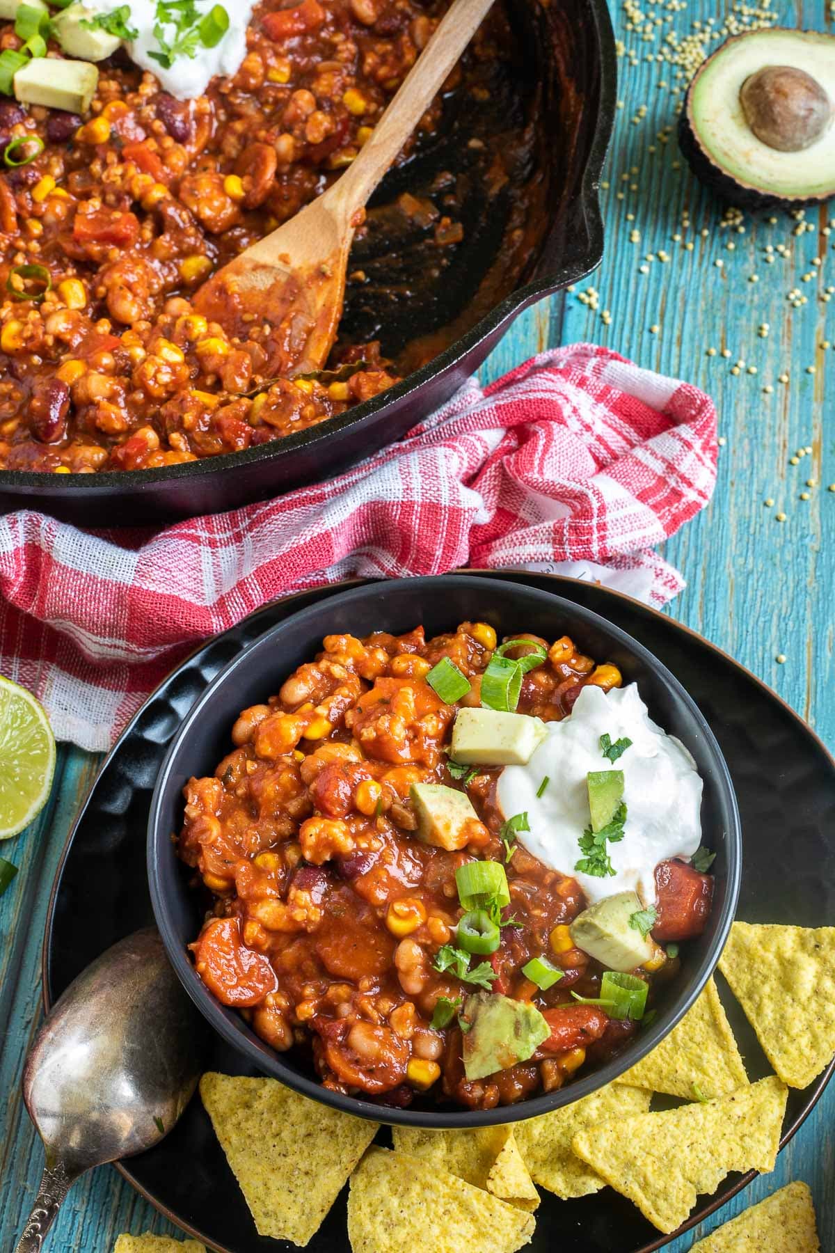 A black bowl with a chunky chili loaded with chopped vegetables and beans, topped with white cream, chopped avocado, and scallion. Leftovers are in a cast iron skillet next to it.