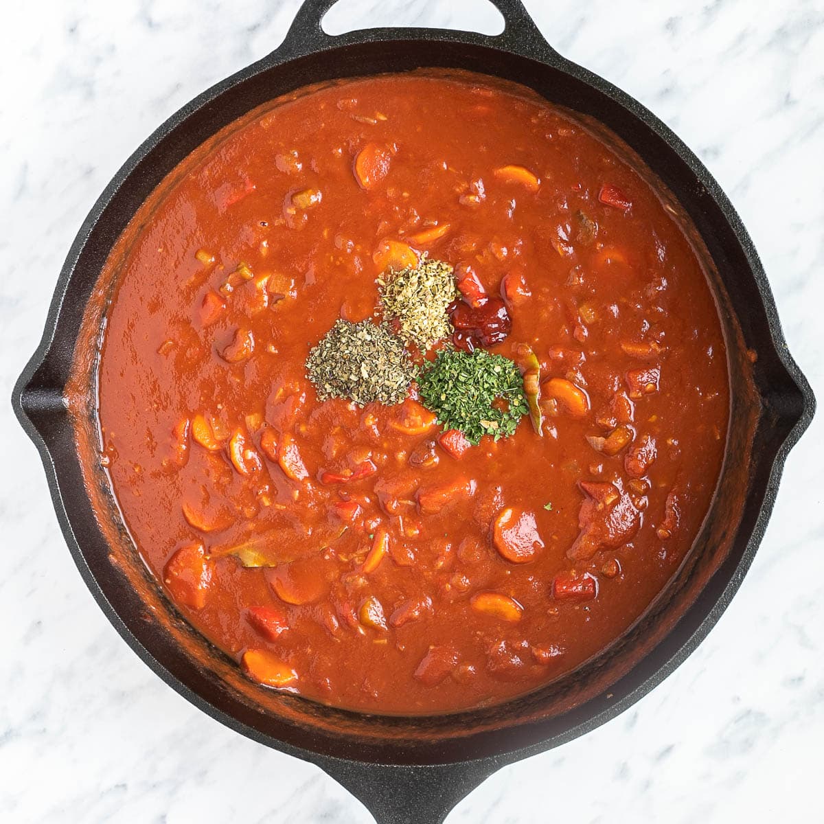 Cast iron skillet with chopped veggies in a thick red tomato sauce topped with 4 heaps of spices.