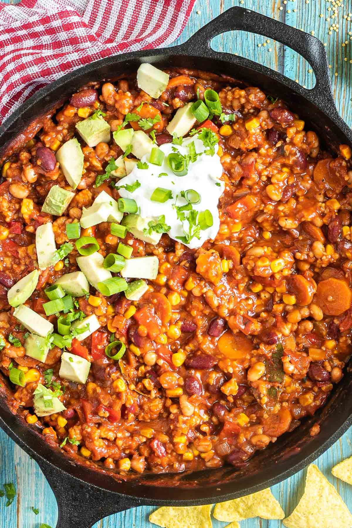 Cast iron skillet with a chunky chili loaded with chopped vegetables and beans, topped with white cream, chopped avocado, and scallion.