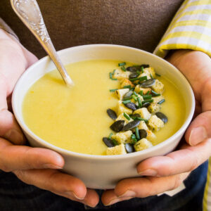 Two hands are holding a white bowl with yellow cream soup topped with croutons, pumpkin seeds and chopped chives.