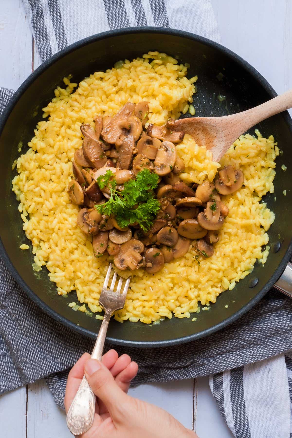 Large black frying pan with yellow creamy rice topped with brown mushroom slices and parsley leaves. A hand is holding a fork and taking some. A wooden spatula is placed in it as well.