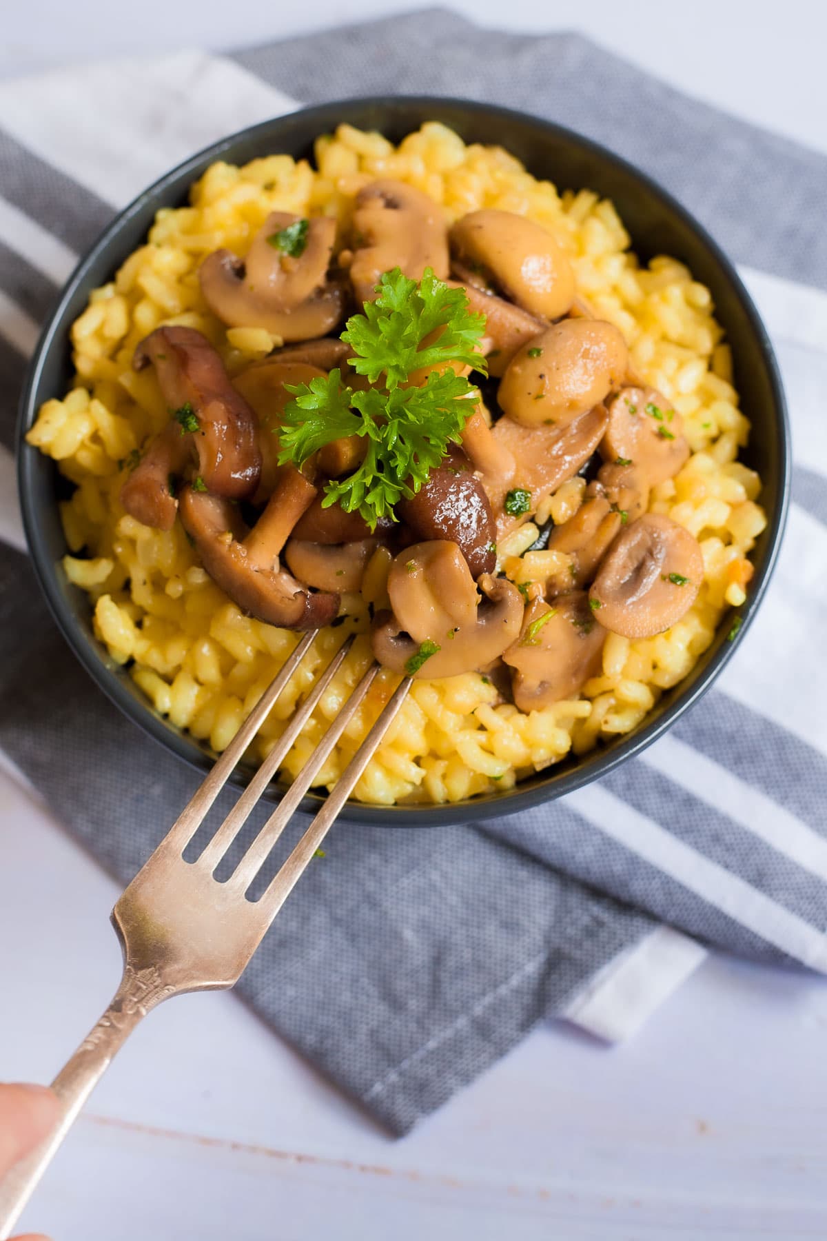 A black bowl of yellow creamy rice topped with brown mushroom slices and green fresh parsley. A fork is placed on top.