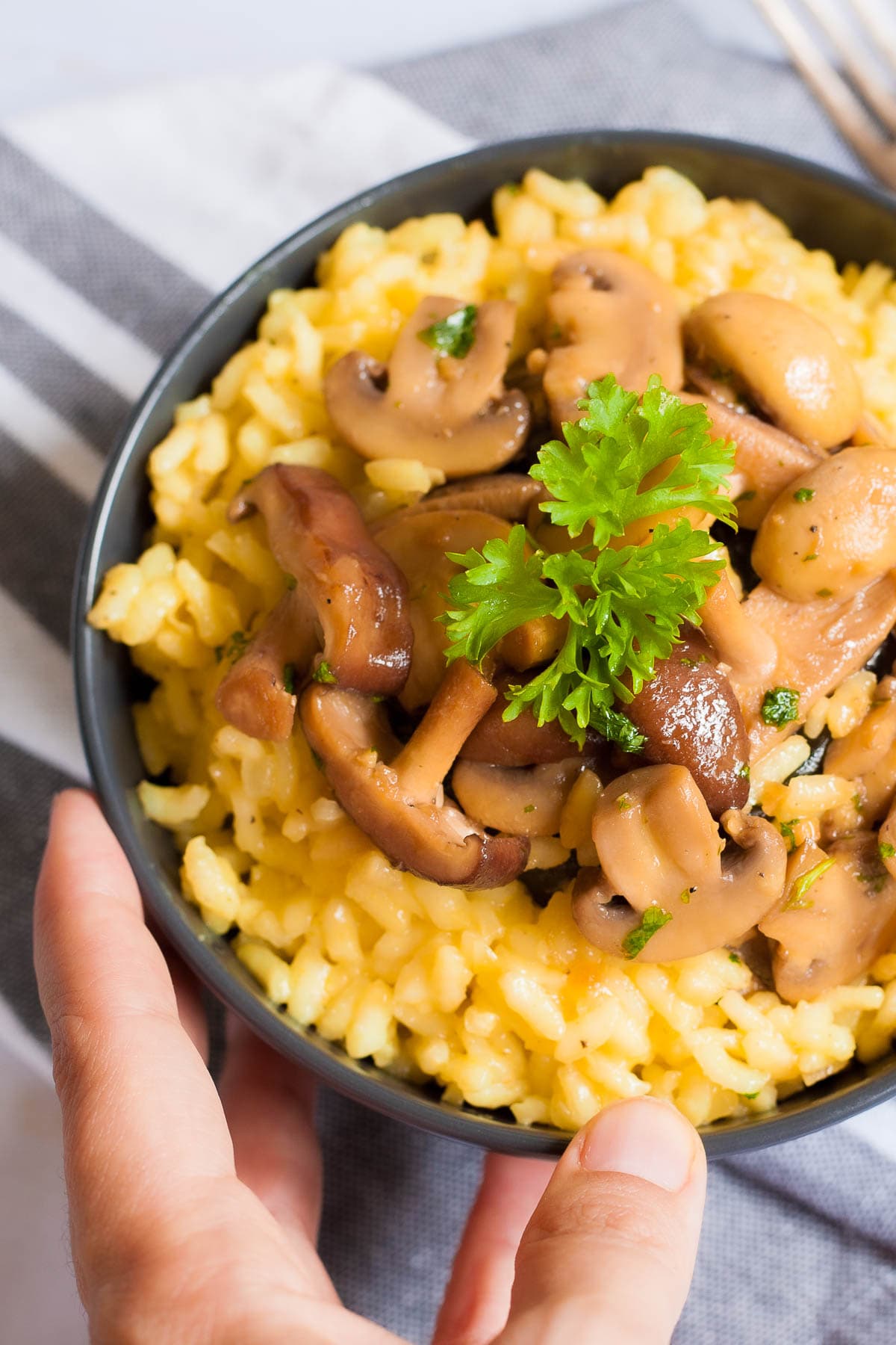 A hand is holding a black bowl of yellow creamy rice topped with brown mushroom slices and green fresh parsley.