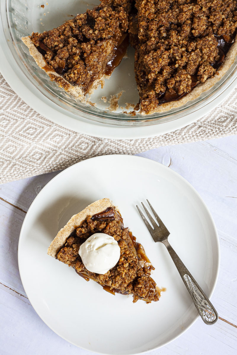 A slice of apple pie on a white plate with brown sauce, apple slices, crumbly topping and a scoop of white vanilla ice cream. There is a fork taking the first bite.