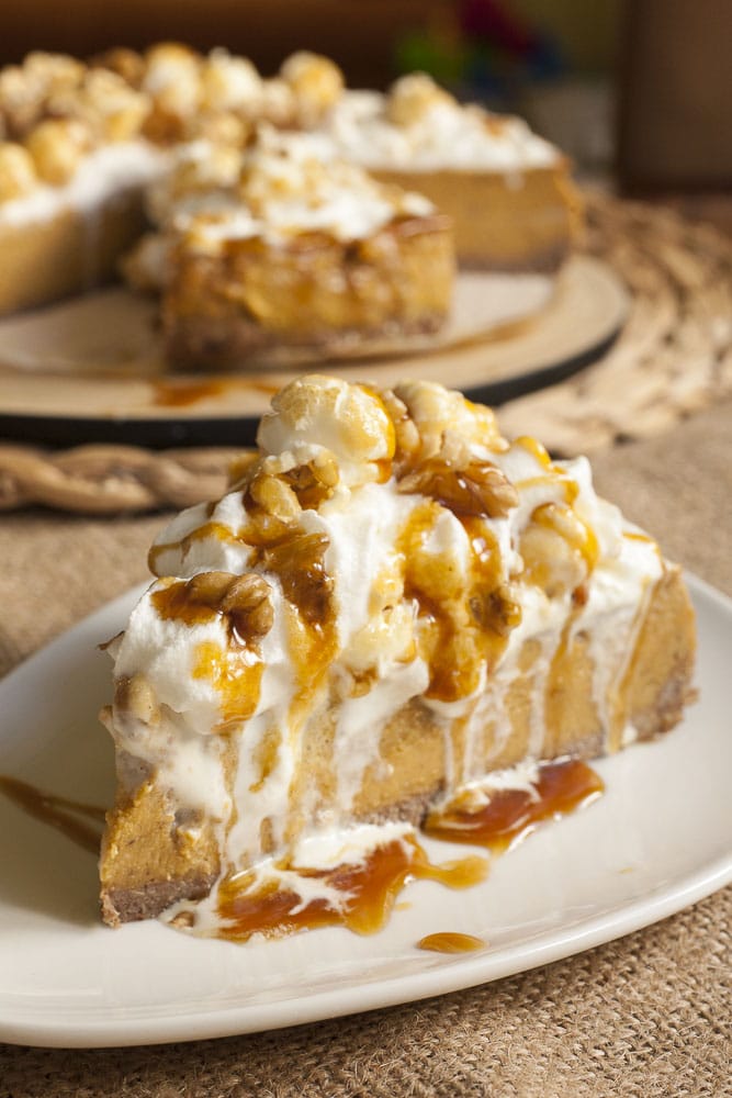 A brown-orange pumpkin colored cake slice on a white plate topped with whipped cream, caramel suace and chopped nuts. 