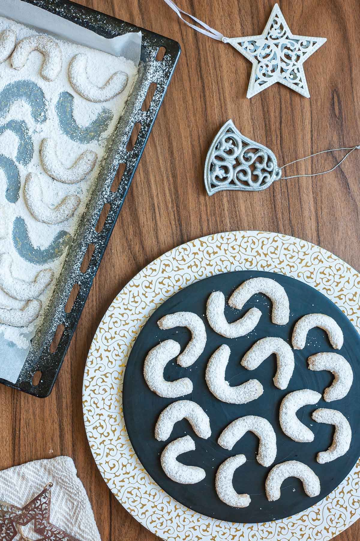 A large stack of crescent cookies on a black plate. Dusted with sugar. A baking tray with the leftover cookies next to it. It is decorated with Christmas ornaments.