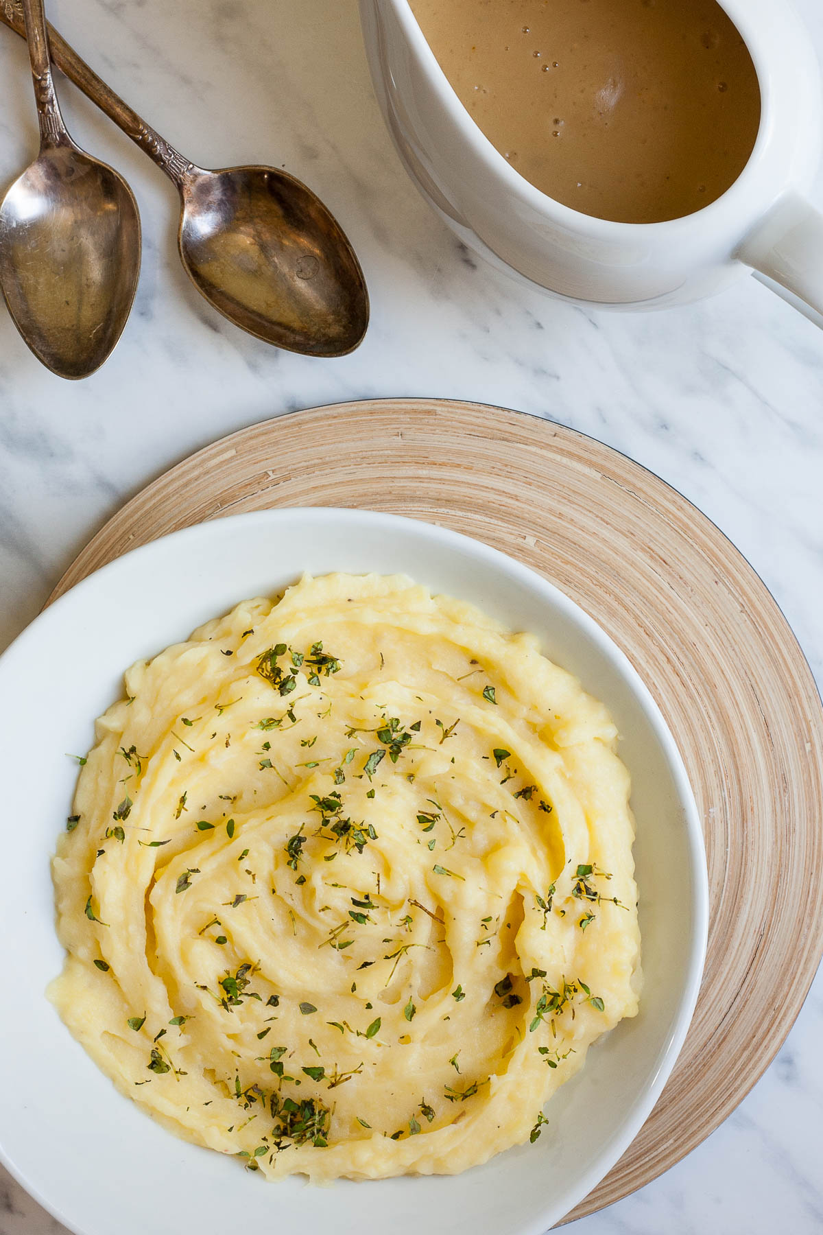 Mashed potatoes sprinkled with green herbs in a white bowl. A small gravy boat with light brown gravy is right behind it.