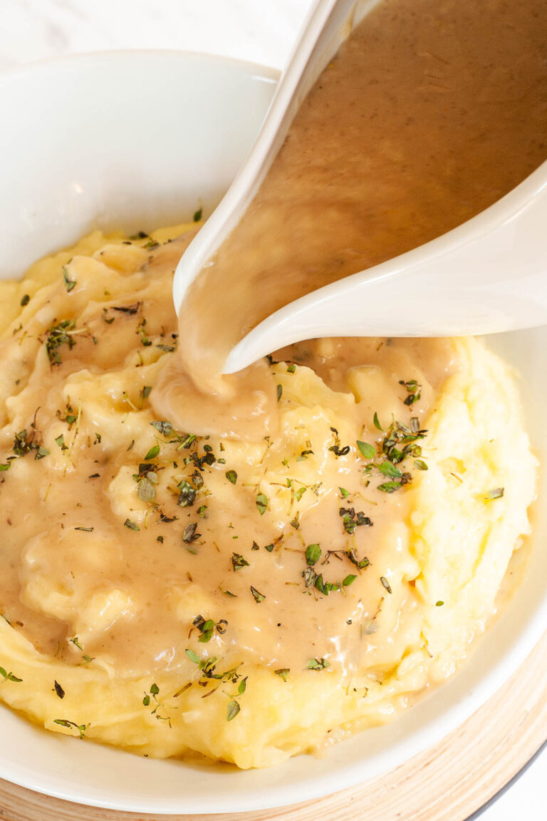 White bowl with mashed potatoes. Light brown gravy is pouring on top of it from a gravy boat.