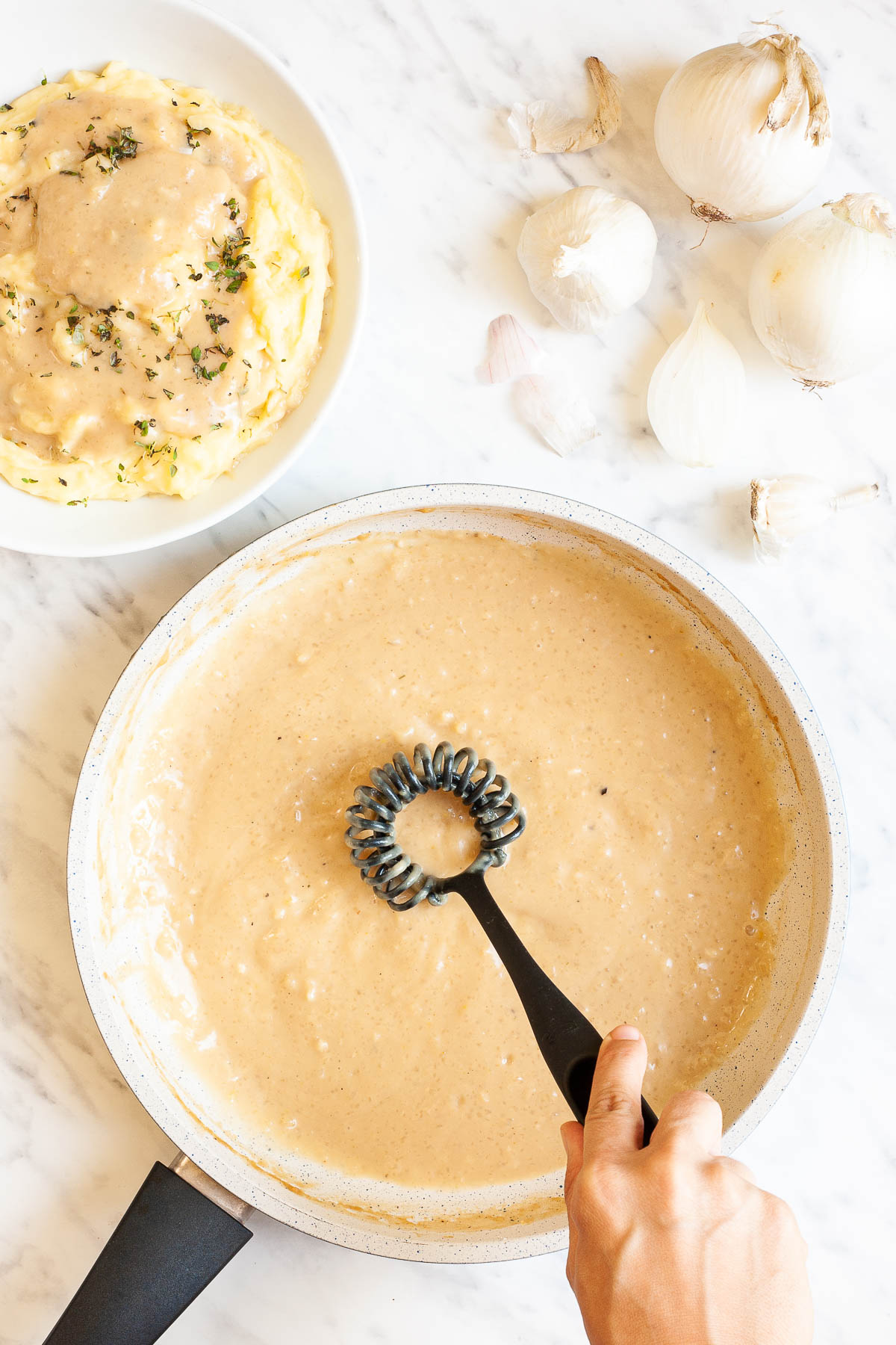 Large white pan with a light brown gravy. A hand is holding a black whisk and whisking the sauce. Mashed potatoes with some gravy on top is next to it as well as some white onions.