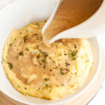 White bowl with mashed potatoes. Light brown gravy is pouring on top of it from a gravy boat.
