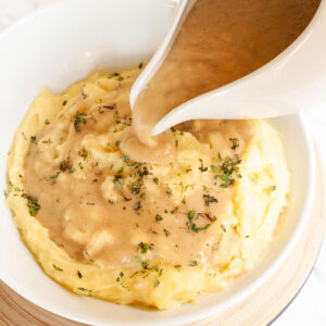 White bowl with mashed potatoes. Light brown gravy is pouring on top of it from a gravy boat.