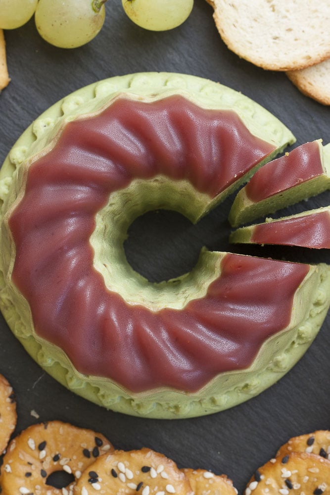 Small green bundt with purple cranberry jelly on top. It is served on a black plate with grapes and crackers. 