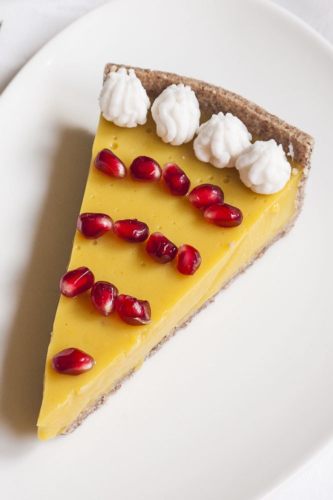 Slice of vegan custard tart on a white plate topped with white whipped cream and pomegranate seeds