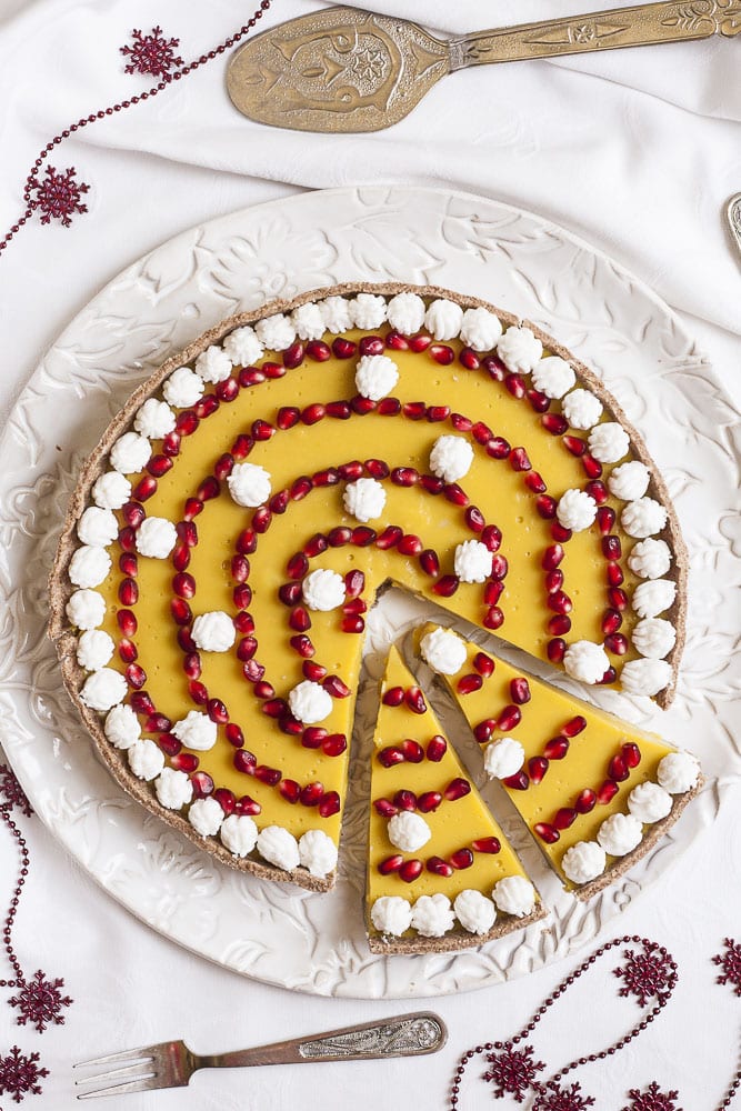 Vegan custard tart topped with pomegranate seeds and coconut whipped cream on a white serving plate. Two slices have been cut. 