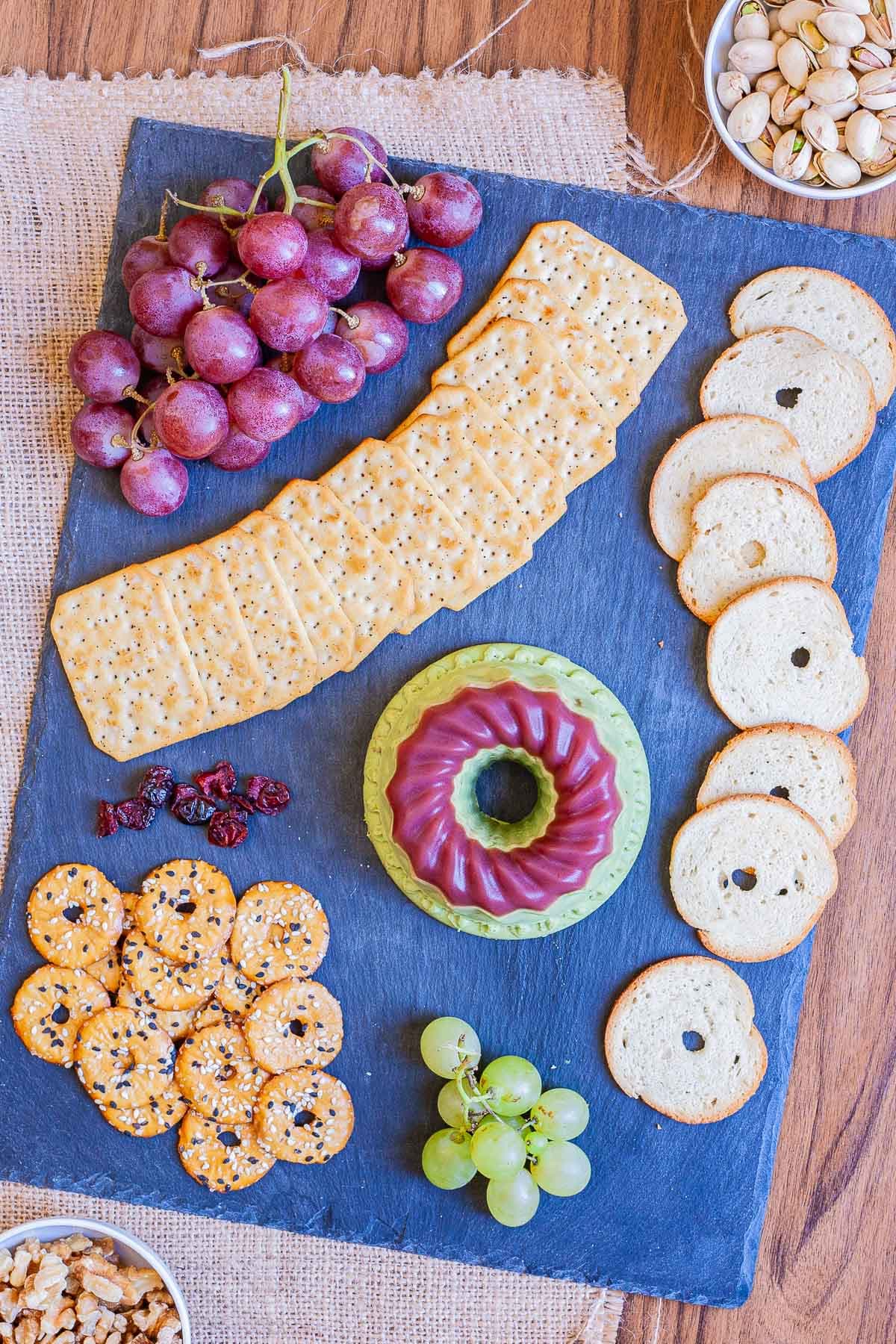 Green cheese shaped as a bundt topped with dark pink jelly. It is surrounded by grapes and different crackers on a black board. 