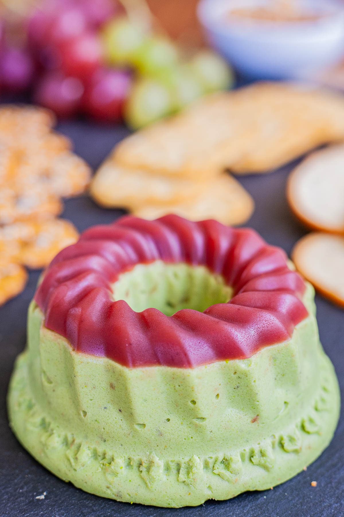 Green cheese shaped as a bundt topped with dark pink jelly. It is surrounded by grapes and different crackers. 