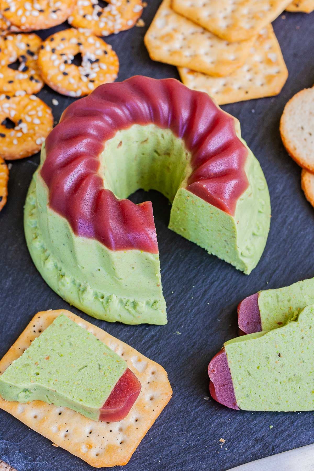 Green cheese shaped as a bundt topped with dark pink jelly. It is surrounded by grapes and different crackers. There is one cracker in the front with one slice of green cheese on it. 