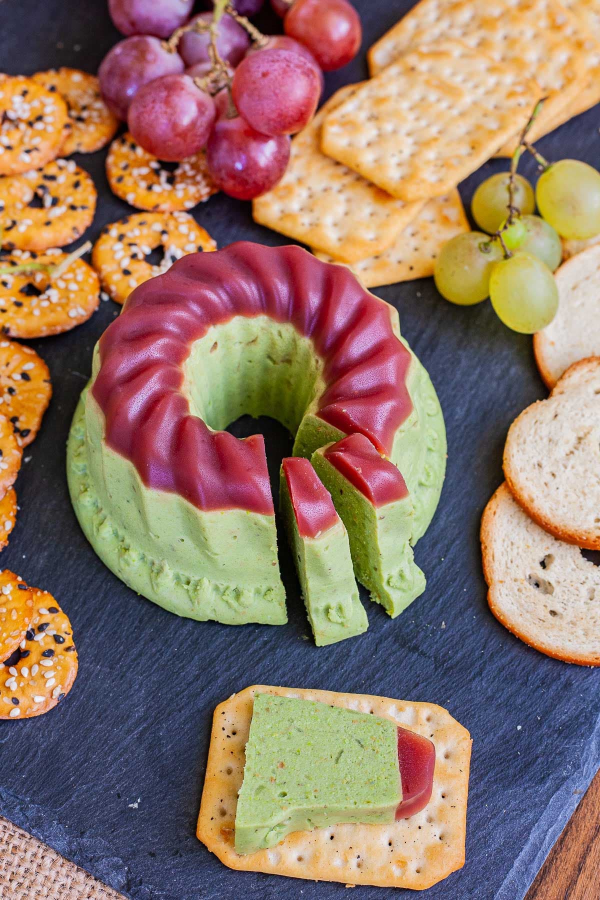 Green cheese shaped as a bundt topped with dark pink jelly. It is surrounded by grapes and different crackers. There is one cracker in the front with one slice of green cheese on it. 