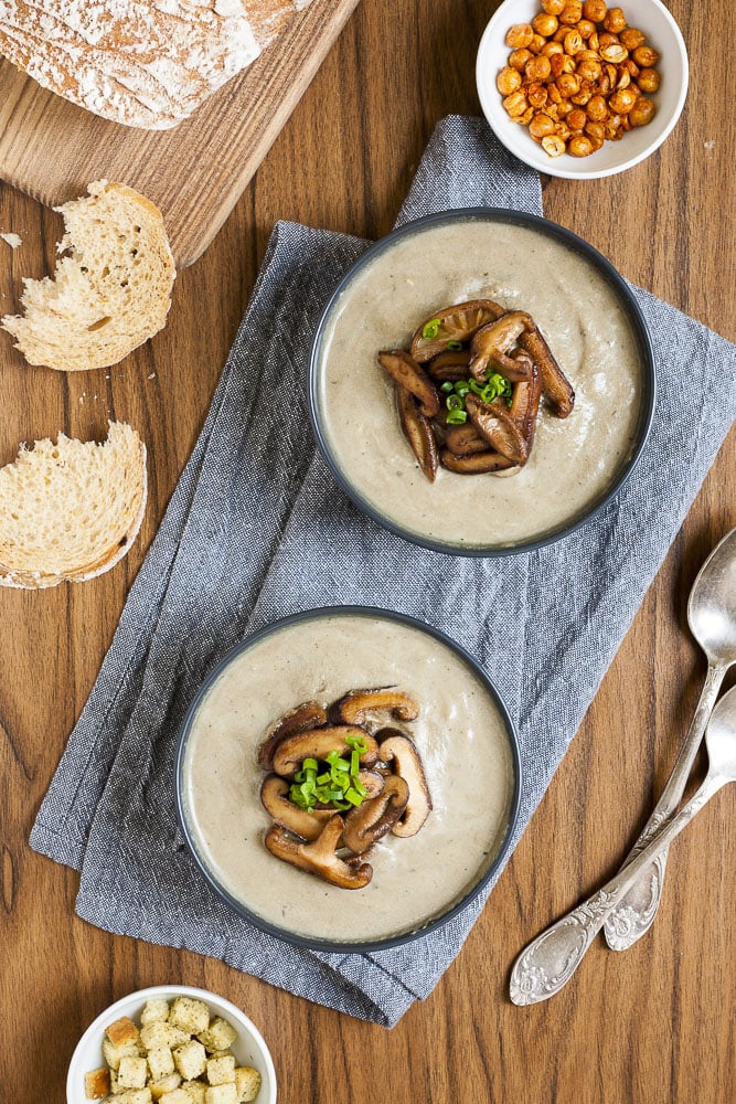 From above Vegan Cream of Mushroom Soup in two black bowls topped with sauteed shiitake mushrooms and spring onion. Croutons and bread slices are around them as garnish. 