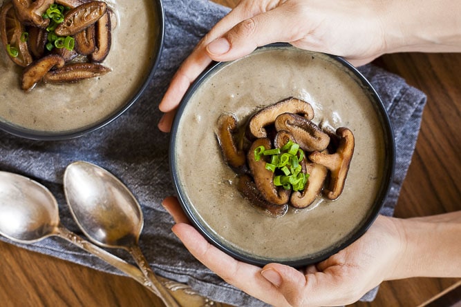 Two hands holding a black bowl of vegan cream of mushroom soup topped with sauteed shiitake mushrooms and spring onion