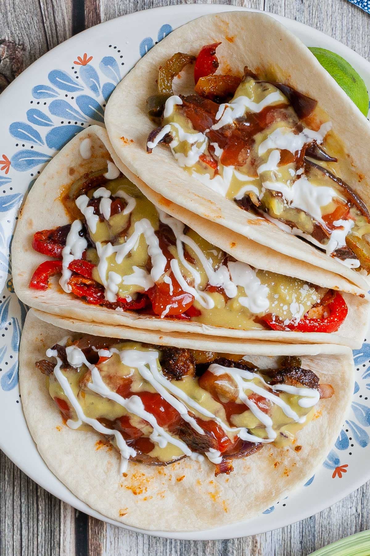 3 soft tortillas folded in half on a white blue plate filled with red pepper slices, purple onion slices and mushroom slices drizzled with white sauce, red salsa and guacamole.