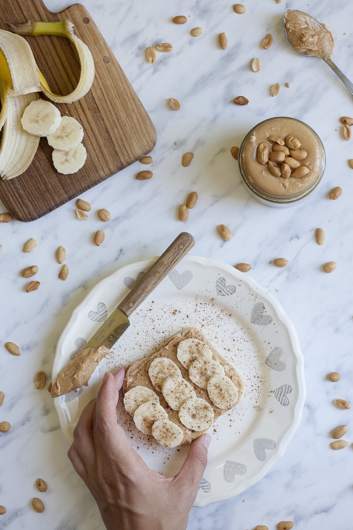 A hand is taking a peanut butter toast covered with banana slices from the plate. A jar of peanut butter, a knife, and some leftover banana slices are around it. 