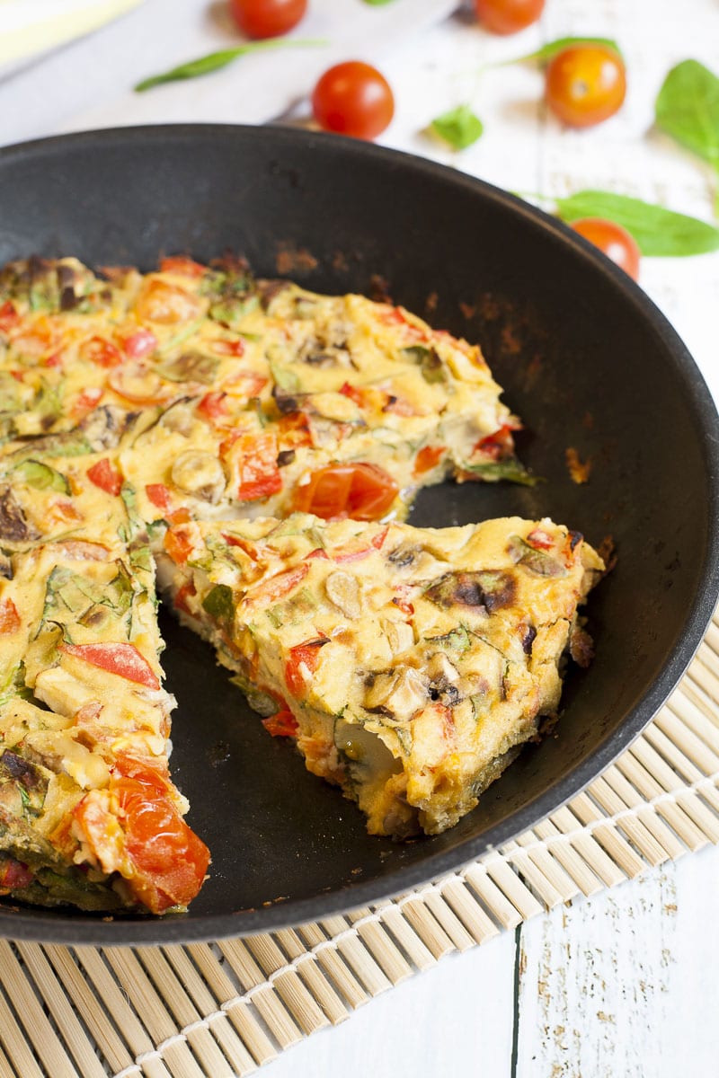 Frying pan with a yellow frittata full of veggies like chopped red bell pepper, green spinach leaves, brown mushroom, red tomatoes. It is already sliced. 