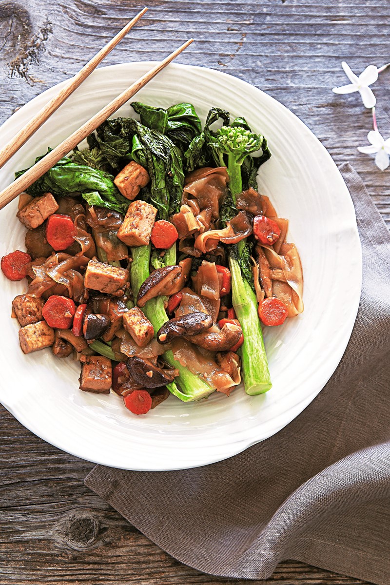 White bowl with brown noodles, bok choy, tofu cubes, carrot slices and purple onion slices. Chopsticks are placed on the side.